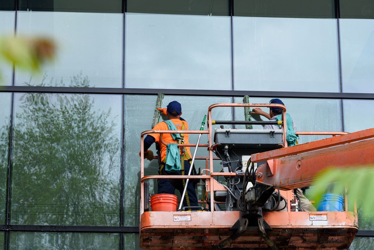 creative-02 Two workers cleaning windows on a glass building facade using a lift platform, enhancing building aesthetics.