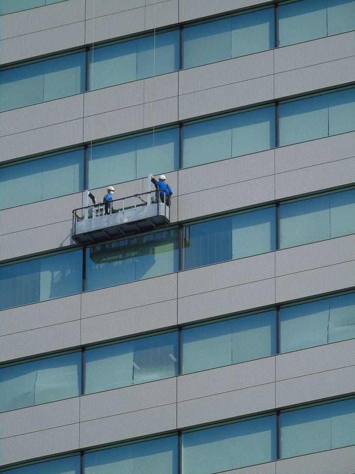 creative-03 Two workers clean windows on a modern glass building facade using a suspended platform.